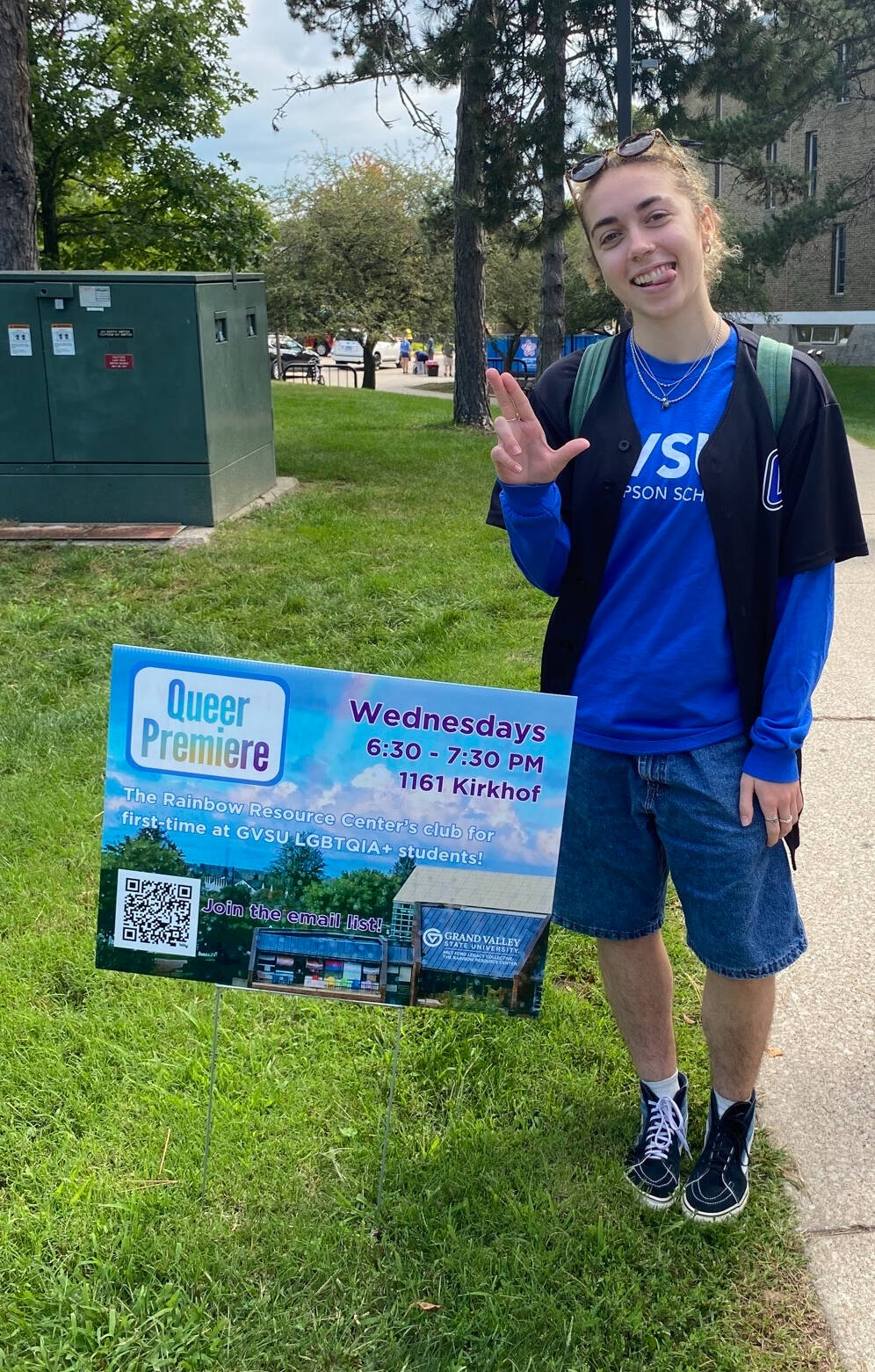 Hannah stands by a Queer Premiere yard sign. She is wearing jean shorts and a baseball tee, and is sticking her tongue out and doing the GVSU anchor up hand sign
